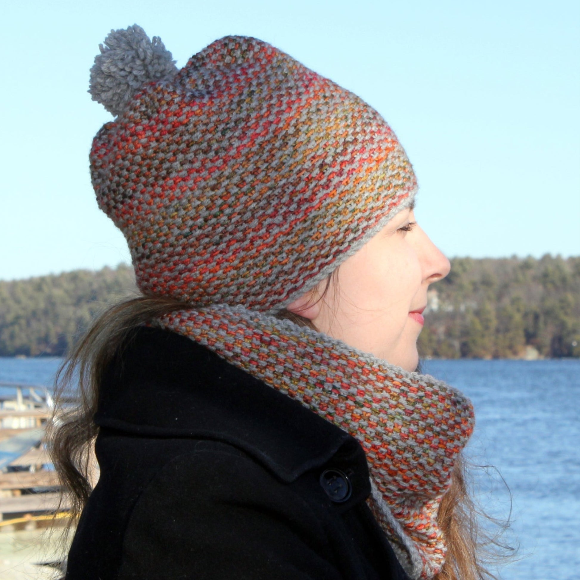 A person wearing the Halcyon Yarn Telephone Line Set, featuring a colorful knit hat with a pom-pom and a matching cowl, stands by a body of water. They are dressed in a dark coat and have long hair. The background includes a wooden dock and a tree-covered shoreline under a clear blue sky.