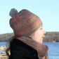 A person in profile view is cozily adorned in the Telephone Line Set, featuring a multicolored knitted hat with a pom-pom and a matching cowl by Halcyon Yarn, both showcasing the classic linen stitch pattern. On a clear day outdoors near a body of water with wooded areas in the backdrop, they are dressed warmly in a dark coat.