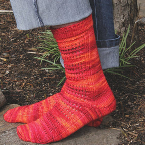 A person wearing the Strings of Rubies Socks from Halcyon Yarn, characterized by their brightly colored, hand-knitted design in shades of red, orange, and yellow using a fingering weight sock pattern, is standing outdoors on a stone path next to some greenery and dark mulch. The person is also wearing light blue jeans that are rolled up at the ankles.