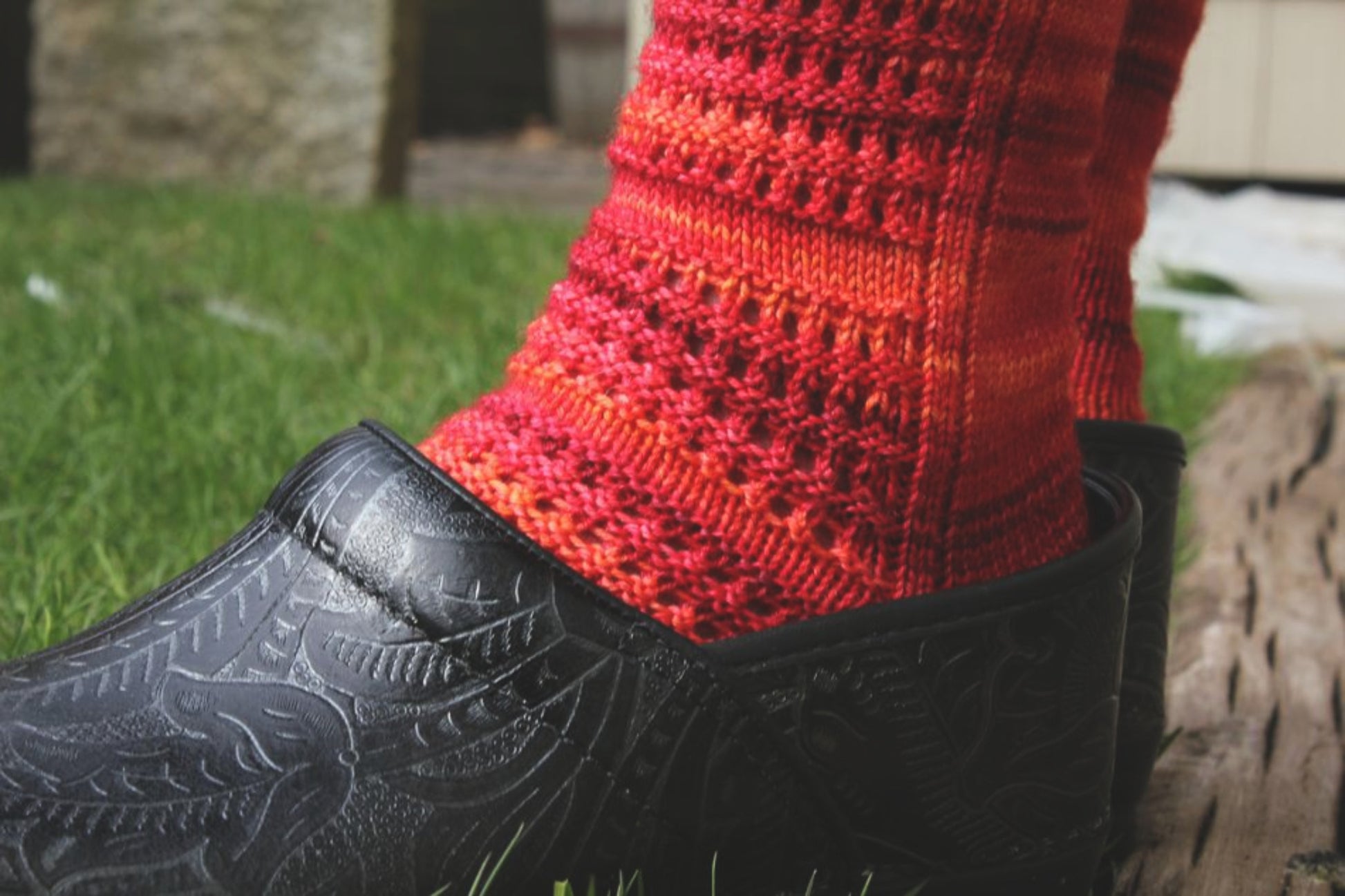 A close-up shot of a foot sporting Halcyon Yarn's Strings of Rubies Socks, showcasing their vibrant red and orange lace-stitch design with a textured pattern. The foot is partially inside an ornately detailed black shoe, resting on green grass with wooden planks in the background.