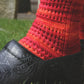 A person is outdoors, wearing the Strings of Rubies Socks from Halcyon Yarn, which are bright red and intricately knitted from sock yarn. They pair these socks with black shoes featuring a detailed embossed design. The person stands on green grass near a piece of weathered wood, showcasing the textured lace pattern they created using circular knitting needles.