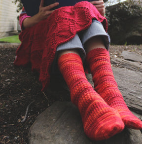 A person sits outdoors with a book, wearing Halcyon Yarn's Strings of Rubies Socks in red and orange women's lace-stitch design. They are cloaked in a red textured blanket, with grey pants peeking out beneath. The background features rocks and grass.