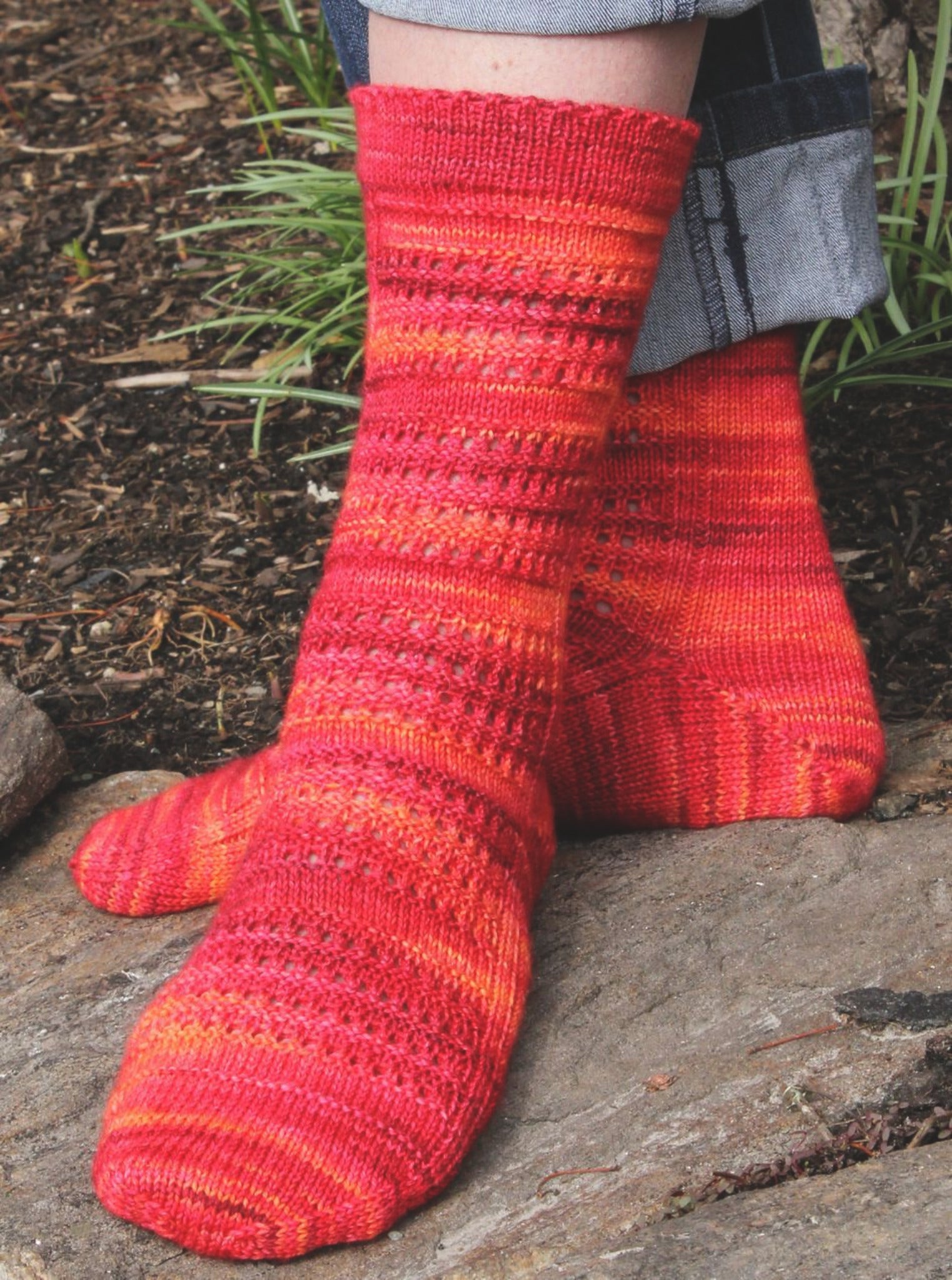 A person wearing the Halcyon Yarn Strings of Rubies Socks, a hand-knitted, multicolored fingering weight sock pattern in shades of red, orange, and pink, stands on a stone surface with one leg crossed over the other. The intricate lace-stitch socks beautifully complement their denim pants. Green plants and soil provide a natural backdrop.