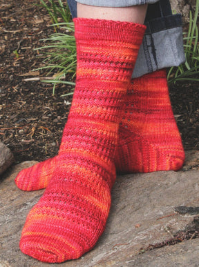 A person wearing the Halcyon Yarn Strings of Rubies Socks, a hand-knitted, multicolored fingering weight sock pattern in shades of red, orange, and pink, stands on a stone surface with one leg crossed over the other. The intricate lace-stitch socks beautifully complement their denim pants. Green plants and soil provide a natural backdrop.