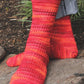 Image of a person wearing the vibrant "Strings of Rubies" socks by Halcyon Yarn, hand-knitted with alternating stripes of red, orange, and yellow hues. The intricate lace pattern adds a textured touch to the sock yarn. The person is standing outdoors on a stone path with some greenery in the background. The cuffs of their jeans are slightly rolled up.