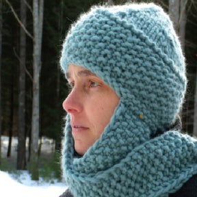 A person with light skin, dressed warmly in The Cuddler, a super bulky knit hat by Halcyon Yarn featuring attached scarves, is outdoors in a forest with snow visible on the ground and trees in the background. They look thoughtful and slightly into the distance.