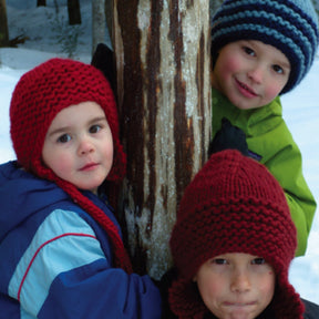 Three children, presumably siblings dressed for winter, gather near a tree trunk. One child in a blue jacket and "The Family Favorite Hat" by Halcyon Yarn in red is in the foreground. Another child in a green jacket and bulky knit hat peeks from behind the tree. A third child, also sporting "The Family Favorite Hat" by Halcyon Yarn made of super soft yarn in red, stands slightly above the first; snow covers the ground.