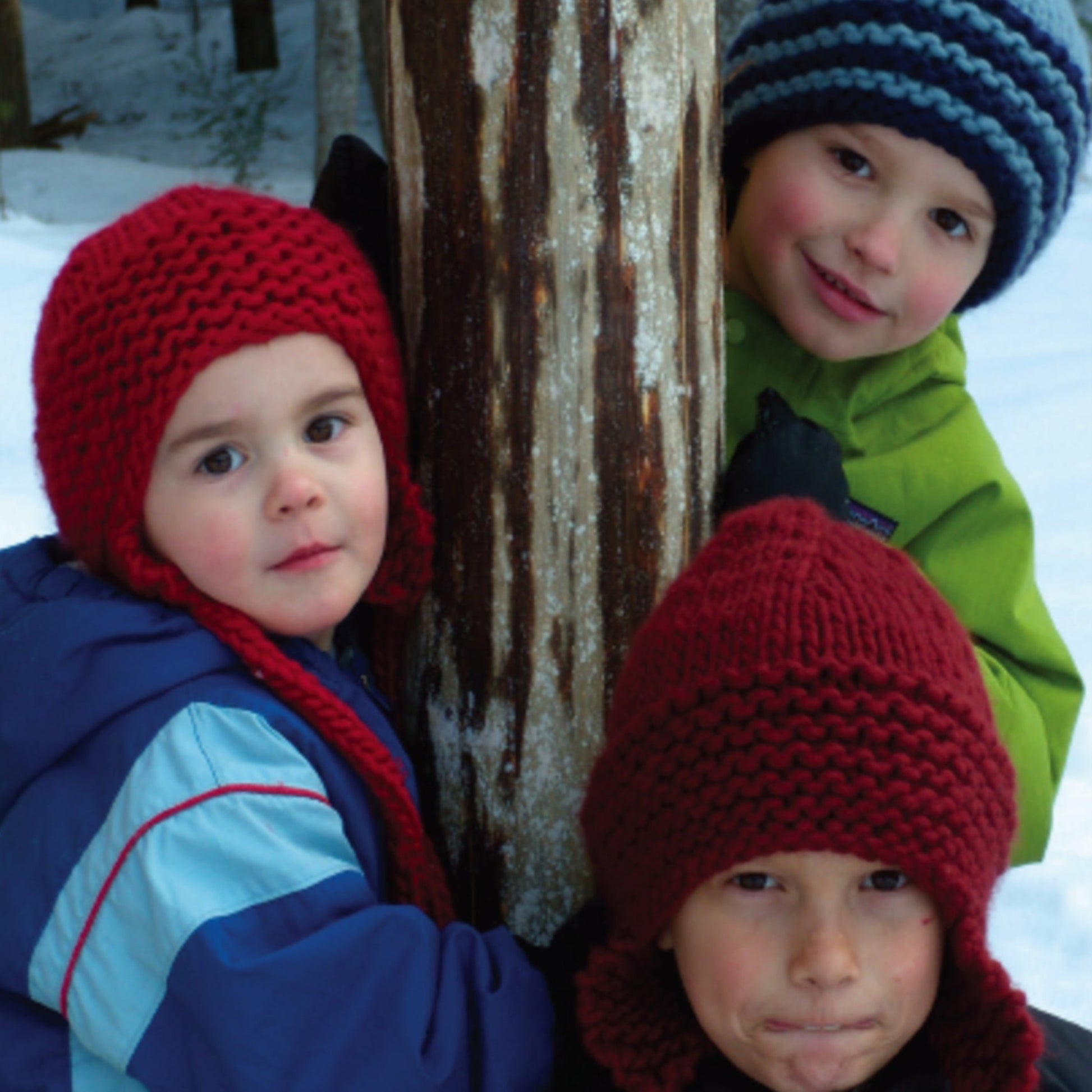 Three children dressed in colorful winter clothing, including The Family Favorite Hat from Halcyon Yarn in vibrant red and blue and green jackets, stand close together near a tree in the snow. They are smiling softly and appear cozy and warm despite the wintry surroundings, capturing a perfect family moment.