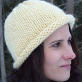 A person with long dark hair, wearing a thick Roll Brim Hat by Halcyon Yarn that exudes classic charm, is shown in profile against a backdrop of trees. The person is looking slightly downward with a neutral expression.