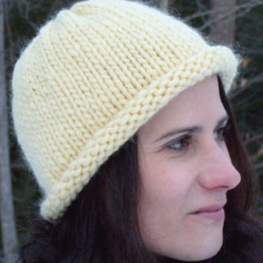 A person with long dark hair is wearing a thick, soft beige Roll Brim Hat from Halcyon Yarn. The background appears to be an outdoor scene with trees. The person is gazing to the right, showing a calm and contemplative expression.