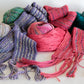 A collection of Scarf Simplicity pieces by Halcyon Yarn, along with assortments of yarn balls, are artfully arranged on a white background. These scarves and yarns showcase various shades of purple, pink, teal, and multicolored hues with intricate knitting patterns. Some scarves feature elegantly fringed ends.