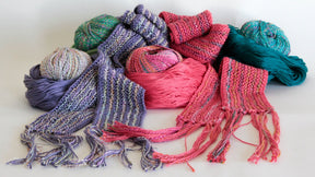 A collection of Scarf Simplicity pieces by Halcyon Yarn, along with assortments of yarn balls, are artfully arranged on a white background. These scarves and yarns showcase various shades of purple, pink, teal, and multicolored hues with intricate knitting patterns. Some scarves feature elegantly fringed ends.