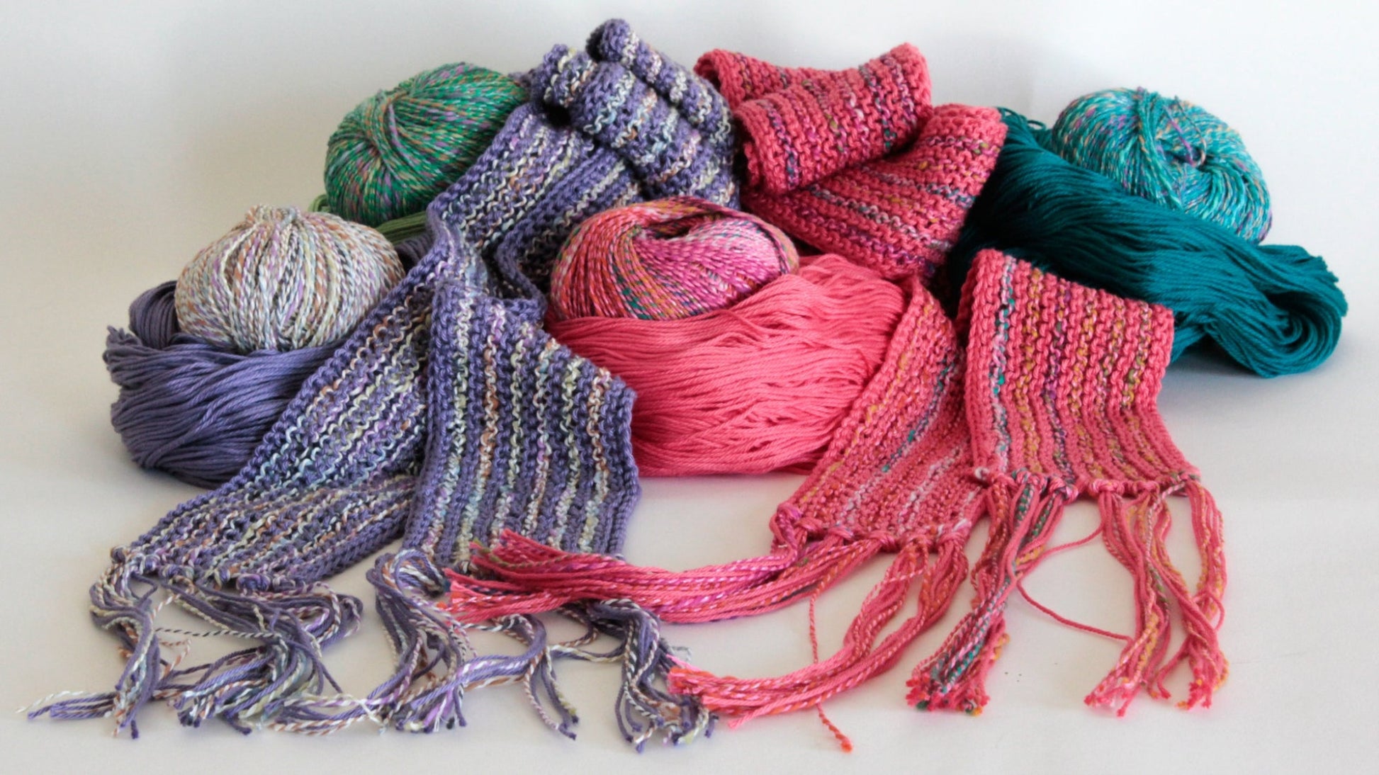 An assortment of Halcyon Yarn's Scarf Simplicity, featuring vibrant striped scarves and skeins of yarn in hues of purple, pink, green, and teal. The distinctively designed scarves are displayed prominently in the foreground with their loose ends showing, while the yarn balls are positioned behind them against a white backdrop.