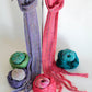 Two "Scarf Simplicity" knitted scarves hang on a stand: one in shades of purple and the other in shades of pink, both products by Halcyon Yarn. Six balls of yarn in various colors (green, blue, purple, multicolored, pink, and teal) are arranged around the base of the scarves, showcasing a vibrant display for any knitting pattern enthusiast.