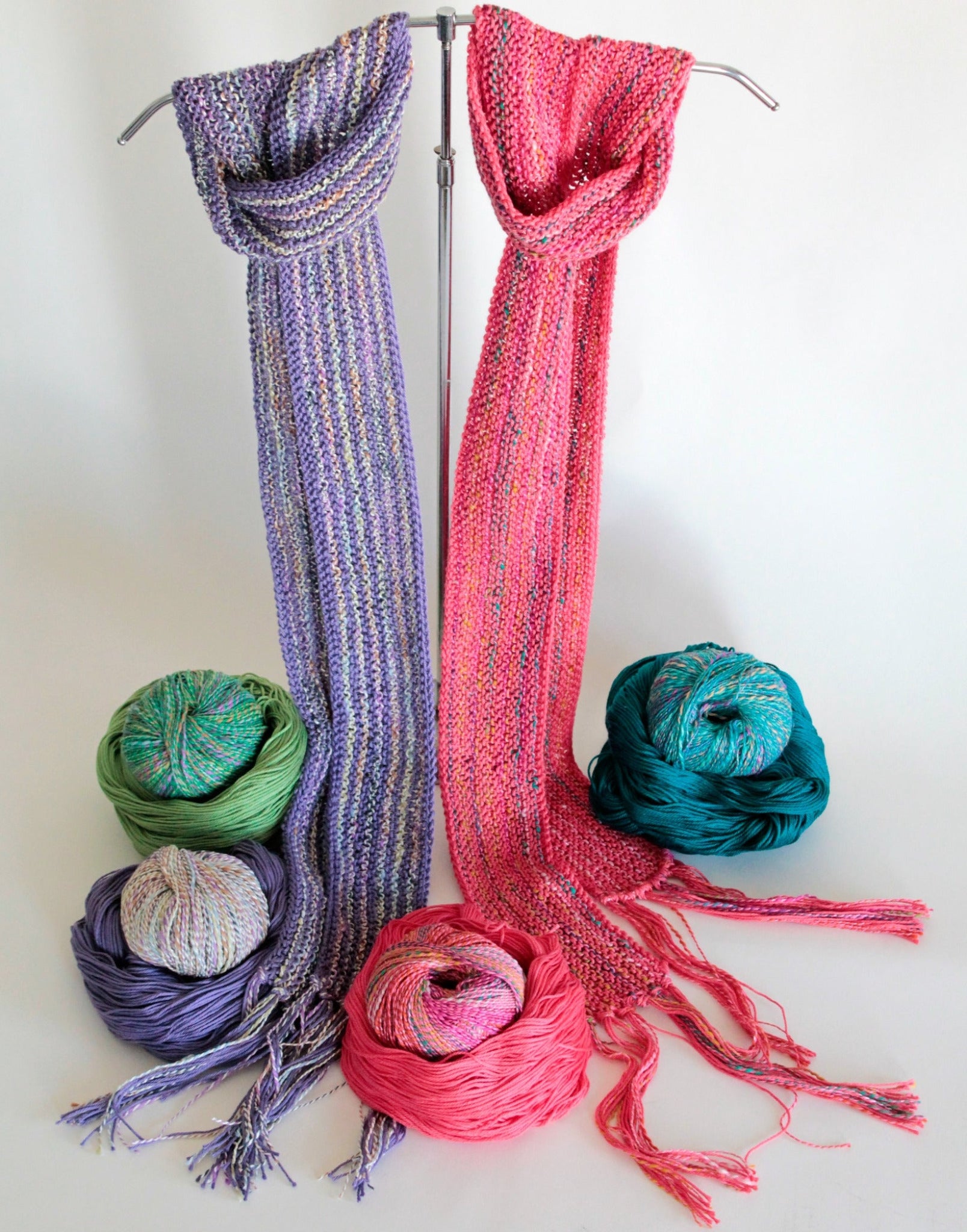 Two "Scarf Simplicity" knitted scarves hang on a stand: one in shades of purple and the other in shades of pink, both products by Halcyon Yarn. Six balls of yarn in various colors (green, blue, purple, multicolored, pink, and teal) are arranged around the base of the scarves, showcasing a vibrant display for any knitting pattern enthusiast.