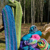 Two Scarf Simplicity knitted scarves by Halcyon Yarn, one in blue and one in green with intricate knitting patterns, hang on a tree trunk in a forest. Below, several balls of yarn in various colors are stacked on a fallen log. Bare trees fill the background, suggesting an early spring or late autumn setting.