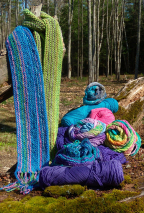 The "Scarf Simplicity, download" from Halcyon Yarn is displayed hanging on a wooden fence in a forest, its blue and green shades showcasing a unique look. Below the scarf, various skeins of yarn in vibrant colors such as blue, pink, purple, and multicolored are arranged on the moss-covered ground.