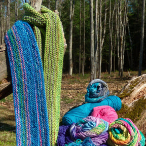 A close-up of colorful yarn and two unique, colorful striped scarves hanging from a branch in a forest clearing. The Scarf Simplicity pieces by Halcyon Yarn are striped in blue and green hues, matching the vibrant balls of yarn in shades of blue, green, pink, and purple resting on the ground.