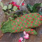 A mitten, crafted using the Snuggly Stuffed Mitten Kit from Halcyon Yarn, features a knitted green design with pink polka dots and rests on a wooden surface surrounded by a bunch of red roses and scattered pink rose petals.