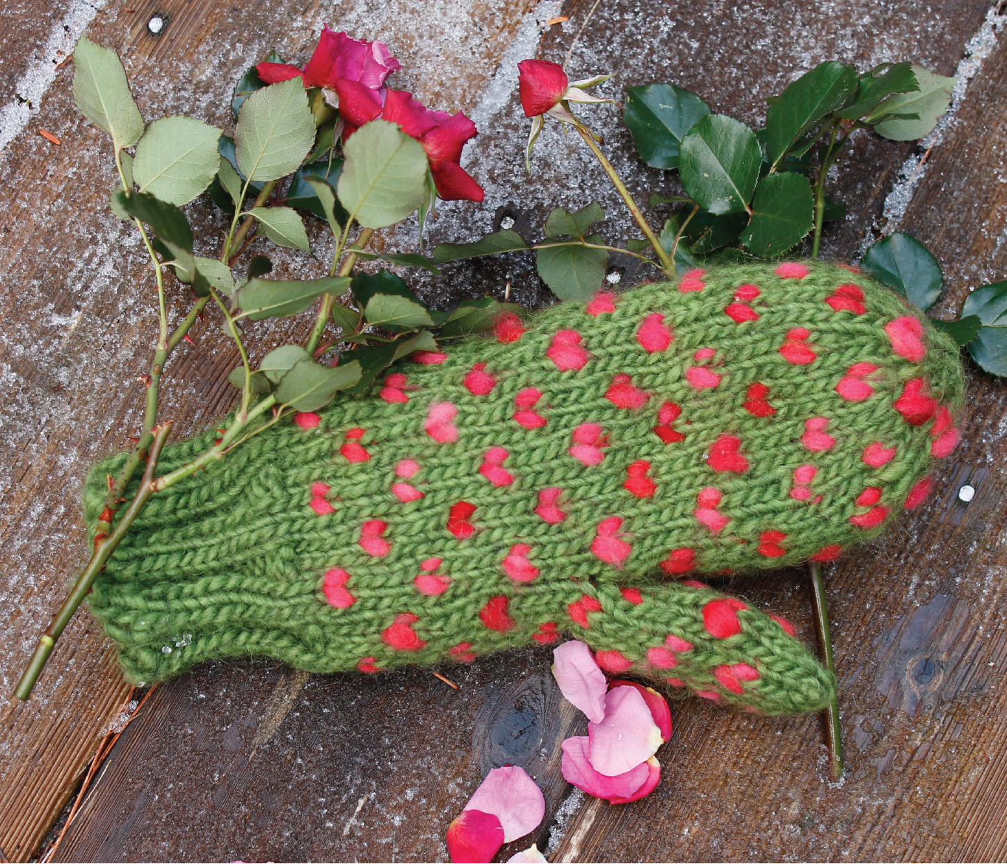 A mitten, crafted using the Snuggly Stuffed Mitten Kit from Halcyon Yarn, features a knitted green design with pink polka dots and rests on a wooden surface surrounded by a bunch of red roses and scattered pink rose petals.