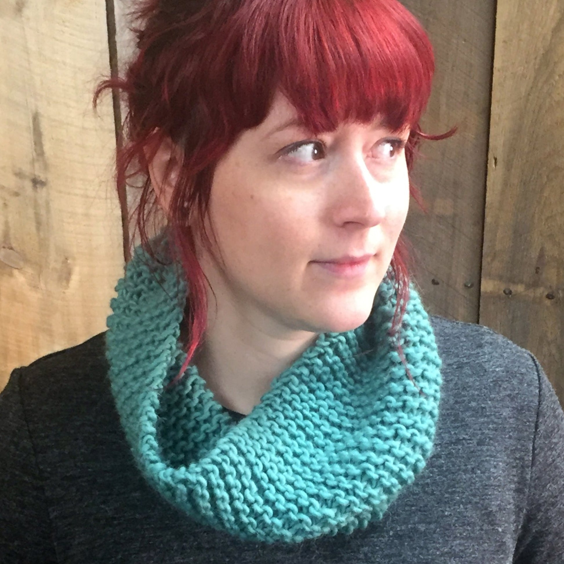 A person with red hair tied back, leaving some bangs and strands loose, is wearing the "Easy: Learn to Knit Cowl" by Halcyon Yarn in teal, made from bulky weight yarn, and looking off to the side. They are dressed in a dark gray top and standing in front of a wooden background.