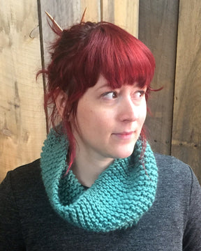 A woman with red hair and bangs wears the "Easy: Learn to Knit Cowl, download" from Halcyon Yarn, knitted in a vibrant teal bulky weight yarn. She pairs it with a dark gray sweater and has knitting needles tucked into her hair. Standing in front of a wooden background, she gazes off to the side.