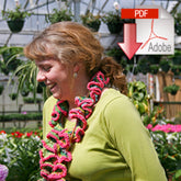 A woman with light brown hair and a green shirt is smiling and wearing a colorful, frilly lei. She is standing in a greenhouse filled with various plants. There is an icon resembling the popular scarf pattern from Halcyon Yarn's "Knit Curlique 2 Ways, download" in the top right corner of the image, featuring a PDF document symbol with a red downward arrow.