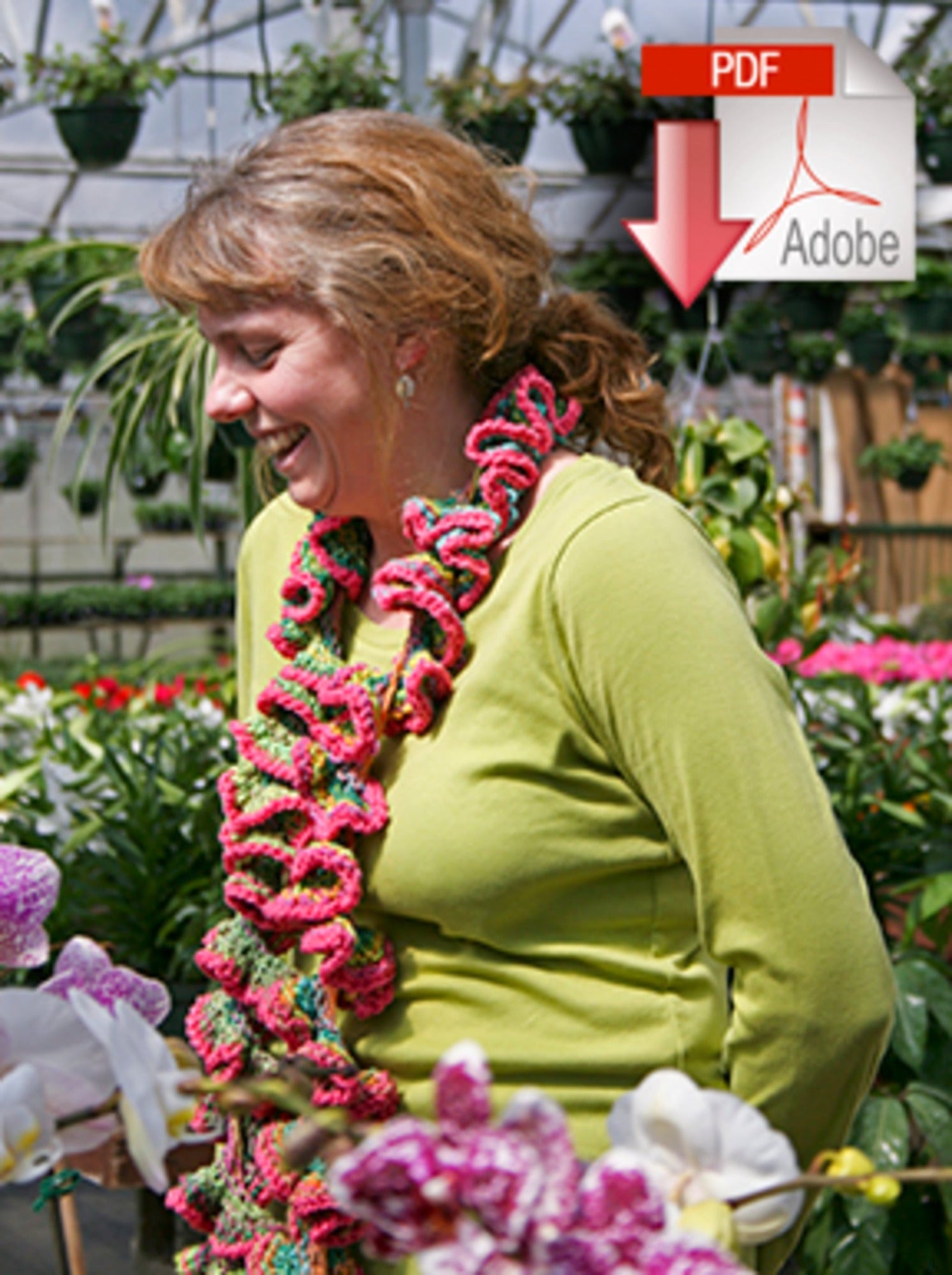 A smiling woman wearing a green shirt and the vibrant, popular Knit Curlique 2 Ways scarf from Halcyon Yarn stands among various flowering plants in a greenhouse. An Adobe PDF icon with a red downward arrow is superimposed in the upper right corner of the image.