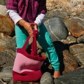 A child wearing a purple sweater and turquoise pants stands on a rocky surface, holding the Shell Seeker Tote by Halcyon Yarn. The sunlit scene captures the lower half of the body as they explore the intricately designed tote—one of those great gifts that spark joy.