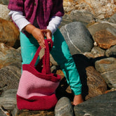 A child wearing a purple sweater and turquoise pants stands on rocky ground, holding a Shell Seeker Tote from Halcyon Yarn. The child is facing downward, their feet slightly bent on the uneven surface, perhaps searching for hidden treasures among the rocks.