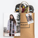 The image displays the Blue Sky Fibers 21 Colors Scarf Kit, which comes in a brown bag filled with vibrant Woolstok yarn balls. A tag illustrates a woman adorned in a gray coat and a multicolored scarf, indicating the scarf kit project. The snowy landscape backdrop conjures up ideas for knitting cozy beanies.