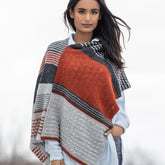 A woman stands outdoors showcasing the Blue Sky Fibers Persimmon Wrap Knitting Kit, which features a multi-colored knit with orange, gray, and black patterns. The wrap is made from Woolstok Light. She has long dark hair and wears a light blue shirt and jeans against a blurred, natural landscape.