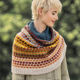 A person with short blonde hair wears a multi-colored knitted shawl over a green dress, standing outdoors in Antelope Valley against a blurred, leafy background. The shawl features intricate patterns in warm hues, reminiscent of the poppy-patterned snood from their favorite Blue Sky Fibers Antelope Valley Snood Knitting Kit by Blue Sky Fibers.