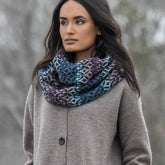 A woman with long dark hair wears a cozy gray coat and the vibrant Blue Sky Fibers North Star Cowl, featuring a colorful, textured checkerboard pattern. She stands outdoors, gazing to the side, against a blurred backdrop of trees.