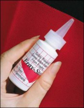 A hand holding a small white squeeze bottle with a pointed tip cap labeled "Fray-Stop" by Yarn Tree, showcasing usage instructions. Ideal for any weaver, it keeps your edges intact with its non-flammable formula that's safe for various materials. The background features red fabric.