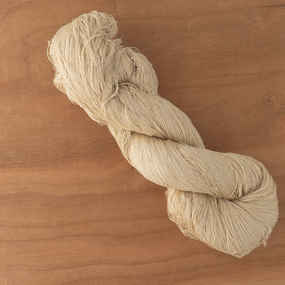 A twisted hank of 2/70 Silk Yarn from Henry's Attic rests on a wooden surface. The smooth and slightly lustrous thread exhibits a light beige color, reminiscent of traditional dyeing techniques used in creating worsted wools.