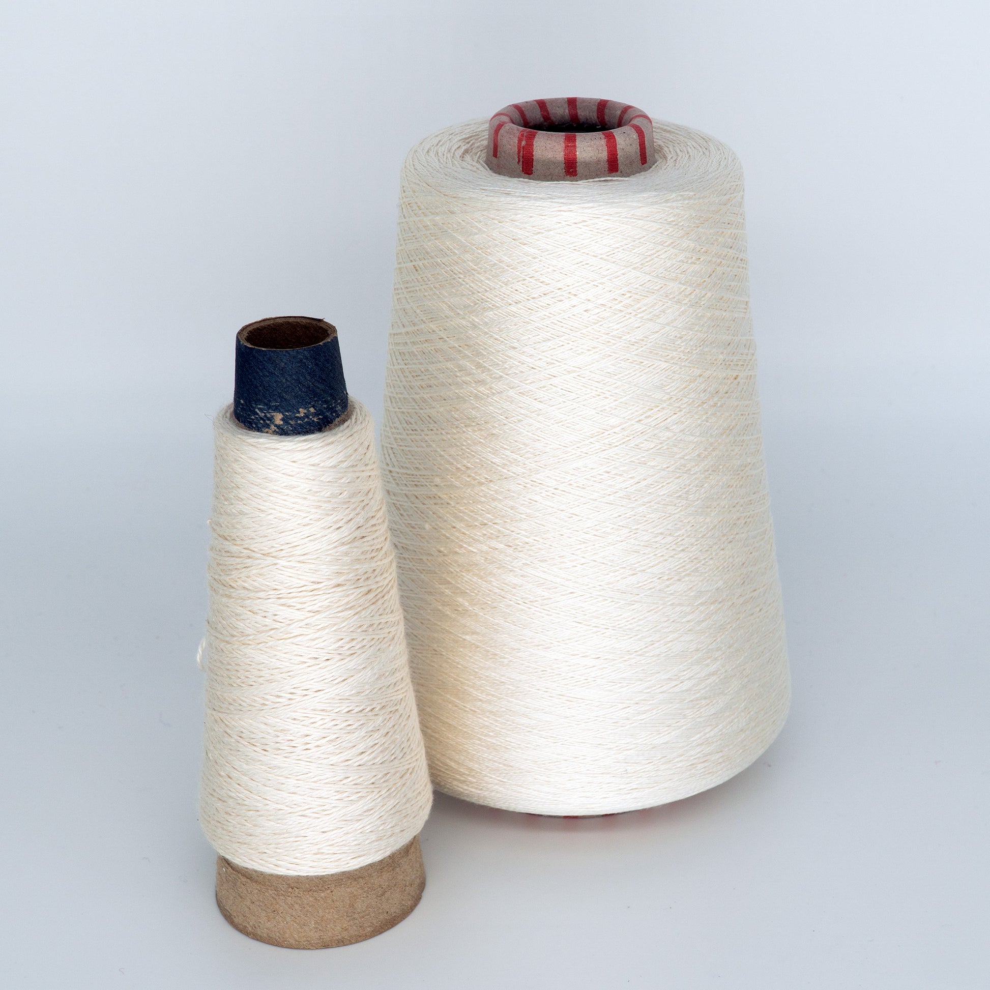 Displayed against a plain background are two spools of Henry's Attic 2/30 Natural Silk Yarn. The larger cone, featuring a red-topped core, is about twice the size of the smaller spool, which has a blue-topped core. Both spools showcase tightly wound off-white yarn, evocative of natural silks.