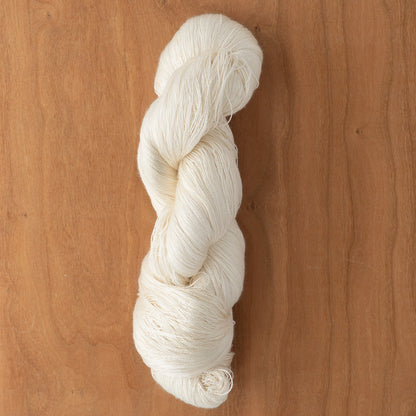 A skein of Henry's Attic 2/30 Natural Silk Yarn is twisted into a neat bundle and placed on a wooden surface. The texture of the off-white yarn is clearly visible, showcasing its soft and slightly fluffy appearance. The wooden background has a natural, light brown tone.