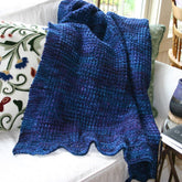 The Family Favorite Throw from Halcyon Yarn, a handwoven, soft and cozy blanket in purple and blue hues, is draped over the back of a white couch. The couch is adorned with a decorative cushion featuring floral embroidery. In the background, there is a wooden chair and a window, along with a table holding a book and a ceramic item.