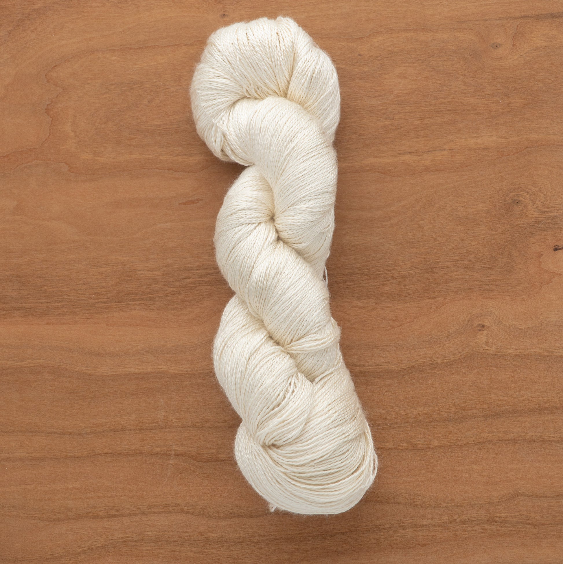 A neatly coiled skein of off-white 2/12 Natural Silk Yarn from Henry's Attic lies on a wooden surface. The silk yarn appears to be soft and finely textured, suitable for knitting or crocheting. The warm tones of the wood contrast with the light color of the yarn, highlighting its delicate fibers.