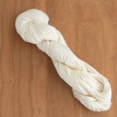 A neatly twisted skein of 2/5 Natural Silk Yarn by Henry's Attic lies on a light wooden surface. The yarn, smooth and well-arranged, highlights its texture and uniform off-white color—ideal for weaving. The wood's grain provides a warm contrast to the soft, creamy hue of the yarn.