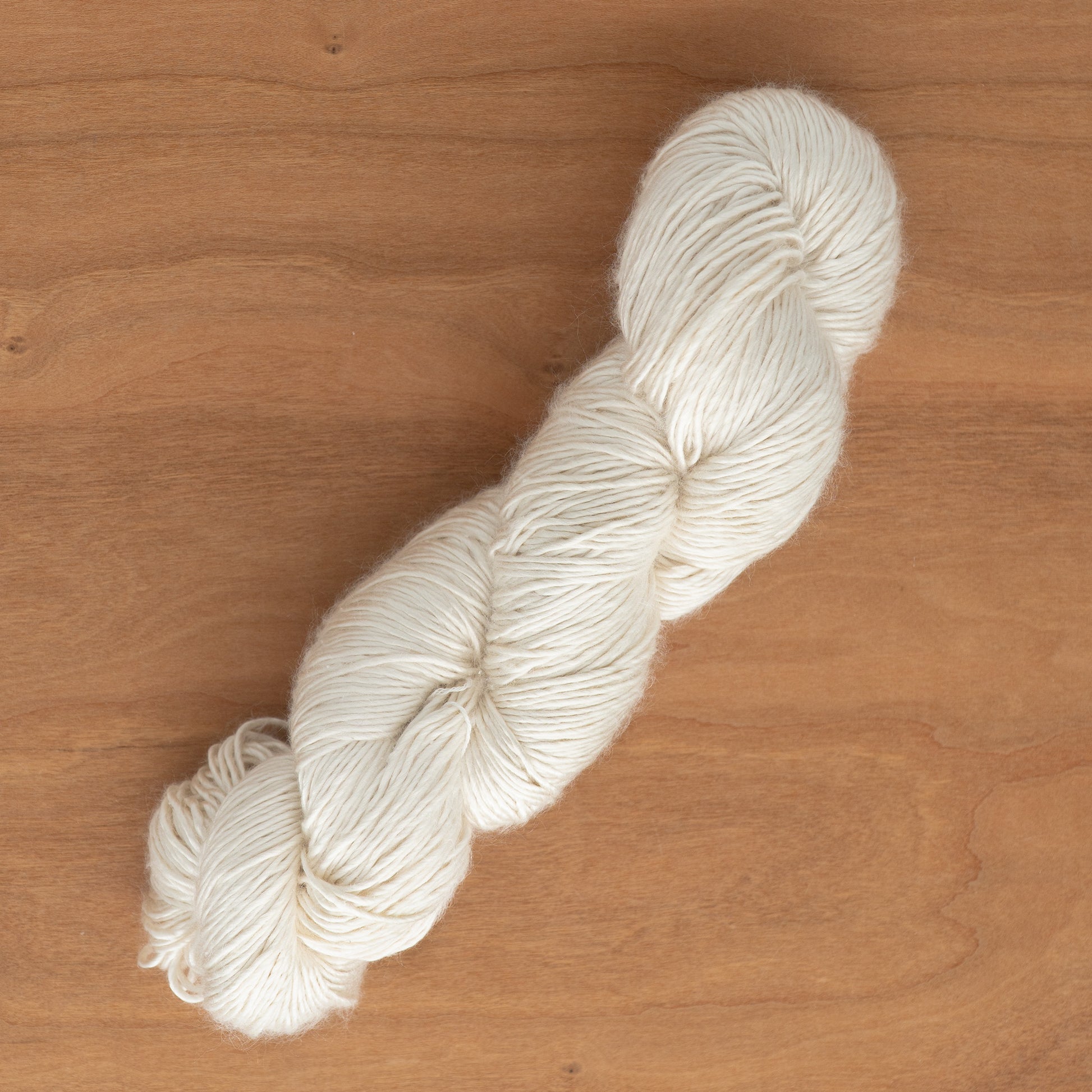 A skein of Soft Twist Natural Silk Yarn by Henry's Attic lies on a wooden surface. The yarn is neatly twisted into a hank, showcasing its soft texture and slightly glossy appearance. This natural fiber yarn hints at an inelasticity that ensures a smooth and cozy finish, suitable for various knitting or crocheting projects.