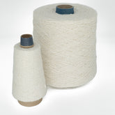 Two cones of cream-colored thread are displayed against a simple white backdrop. The larger cone is cylindrical with a blue center, and the smaller one has a conical shape with a brown base and also features a blue center. Both cones, identified as 2-Ply Silk Noil Yarn (Raw Silk) from Henry's Attic, are tightly wound with thick thread, making them perfect for textured stitches.