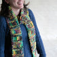 A person with shoulder-length dark hair and a big smile is wearing a blue knitted sweater and a colorful, chunky, hand-knitted Drop Stitch Scarf by Halcyon Yarn that looks incredibly warm. They are standing against a plain background.