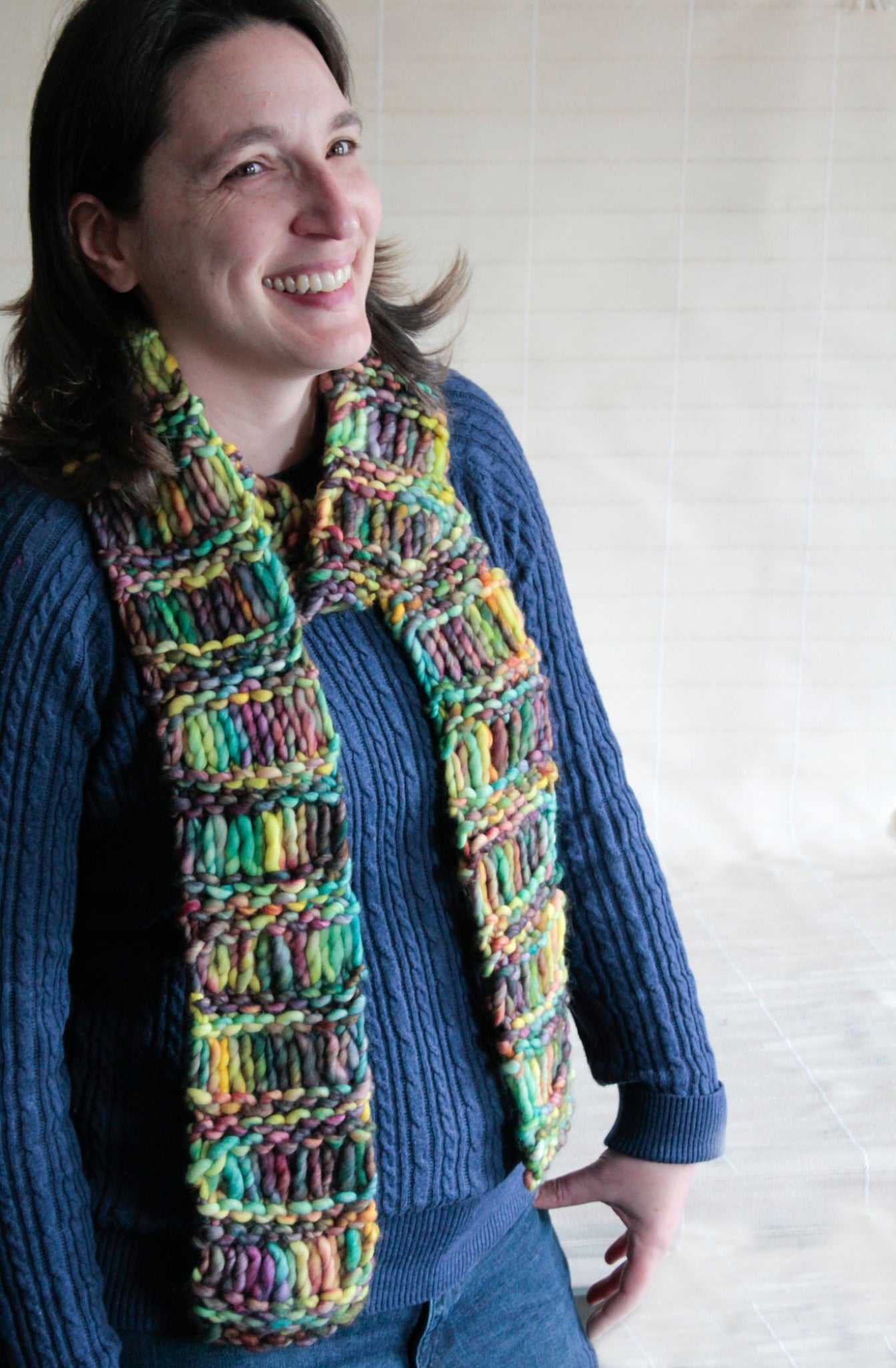 A person with shoulder-length dark hair and a big smile is wearing a blue knitted sweater and a colorful, chunky, hand-knitted Drop Stitch Scarf by Halcyon Yarn that looks incredibly warm. They are standing against a plain background.