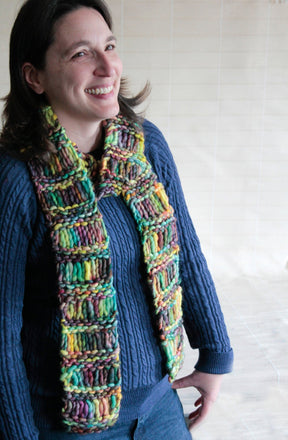 A person wearing a blue cable-knit sweater is smiling while showcasing the Halcyon Yarn Drop Stitch Scarf, download. The warm, multicolored chunky knit scarf features a vibrant mix of yellow, green, and purple. The background is a plain, light-colored wall.