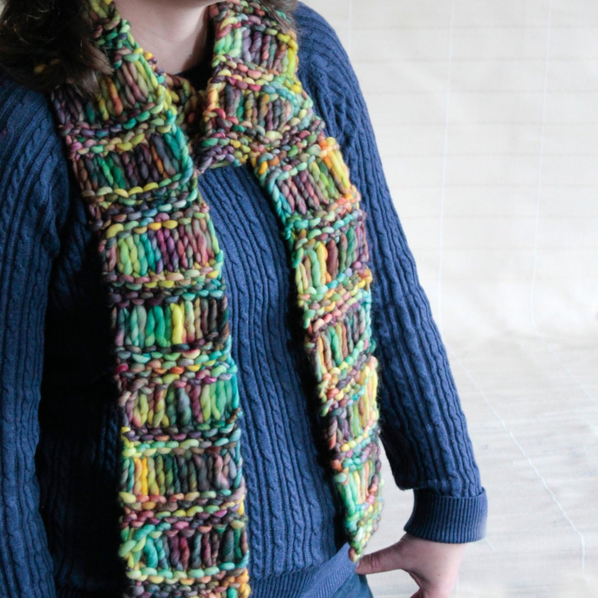 A person wearing a blue sweater dons the warm, multi-colored Halcyon Yarn Drop Stitch Scarf with a rectangular pattern. The background is a neutral off-white color, highlighting the cozy scarf's blend of green, yellow, red, and purple hues.