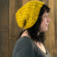 A person with wavy dark hair is wearing a large Monolith Hat, a yellow crocheted beanie from Halcyon Yarn made with bulky weight yarn, along with a dark gray shirt. They are standing in profile against a wooden wall background.