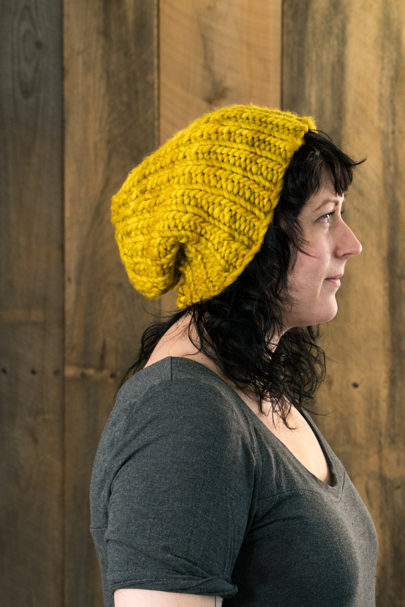 A person with wavy dark hair is wearing a large Monolith Hat, a yellow crocheted beanie from Halcyon Yarn made with bulky weight yarn, along with a dark gray shirt. They are standing in profile against a wooden wall background.