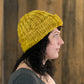 A person with medium-length dark hair is seen in profile, wearing the Monolith Hat from Halcyon Yarn. The chunky, hand-knitted yellow beanie is crafted from bulky weight yarn. They have a neutral expression and are standing in front of a wooden backdrop, alluding to the quick and easy creation made with US 11 and US 15 knitting needles.